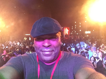 Mickey is a Black man, wearing a flat cap and grey t-shirt and a lanyard. He is taking a selfie in front of a big crowd at a club